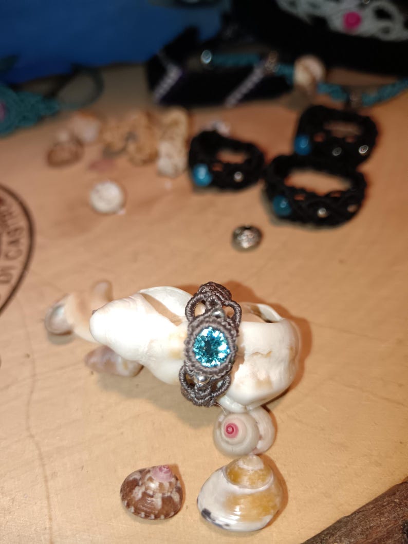 May include: A handmade macrame ring with a teal-colored gemstone set in a detailed, woven design. The ring is displayed on a white seashell, with other seashells and macrame bracelets in the background. The ring's intricate knotwork and vibrant stone are the focal points.