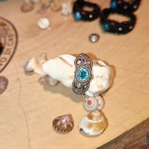 May include: A handmade macrame ring with a light blue gemstone center, displayed on a seashell. The ring is brown and intricately woven. Other macrame bracelets with blue accents are in the background. The setting is a wooden surface with seashells.