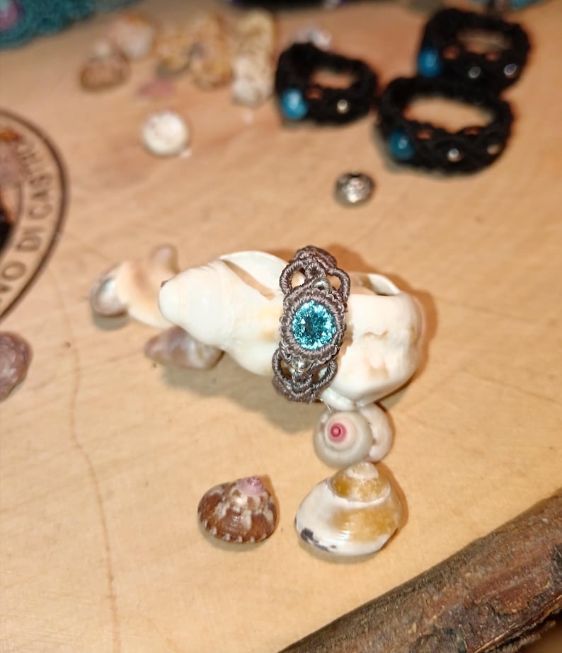 May include: A handmade macrame ring with a light blue gemstone center, displayed on a seashell. The ring is brown and intricately woven. Other macrame bracelets with blue accents are in the background. The setting is a wooden surface with seashells.