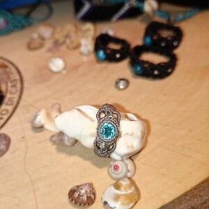 May include: A handcrafted macrame ring with a teal-colored gemstone, displayed on a seashell. The ring features intricate knotting and is surrounded by other seashell accents and jewelry pieces, creating a beachy aesthetic.