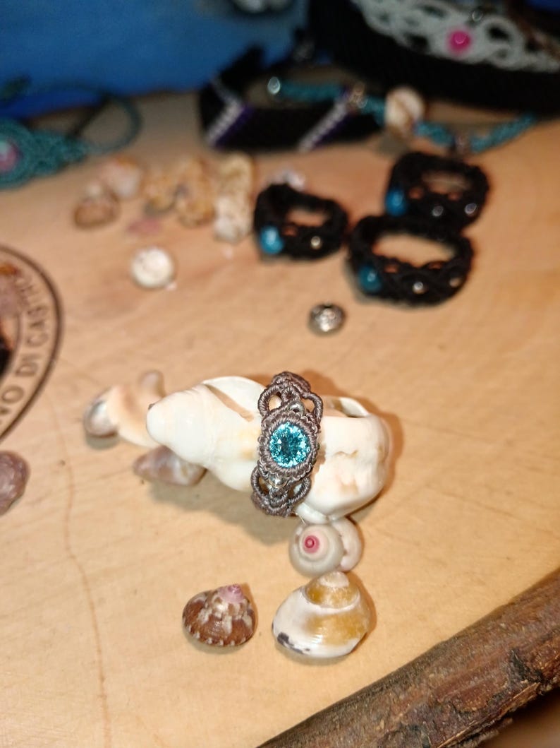 May include: A handcrafted macrame ring with a teal-colored gemstone, displayed on a seashell. The ring features intricate knotting and is surrounded by other seashell accents and jewelry pieces, creating a beachy aesthetic.