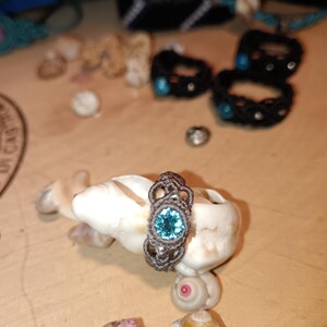 May include: A handmade macrame ring with a teal-colored gemstone set in a decorative, openwork design. The ring is displayed on a white seashell, with other seashells and macrame bracelets in the background. The ring's intricate knotwork and vibrant stone create a unique, bohemian aesthetic.
