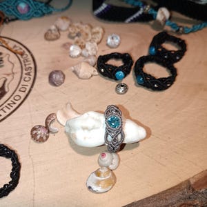 May include: A collection of handmade jewelry, including macrame rings with turquoise accents and bracelets. A silver-toned macrame ring with a blue gemstone sits on a seashell, surrounded by other shells and beads.