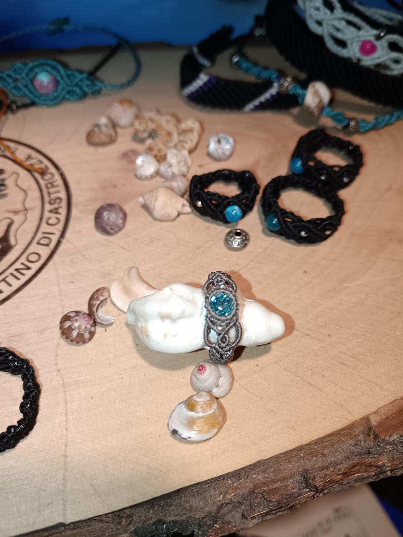May include: A collection of handmade jewelry, including macrame rings with turquoise accents and bracelets. A silver-toned macrame ring with a blue gemstone sits on a seashell, surrounded by other shells and beads.
