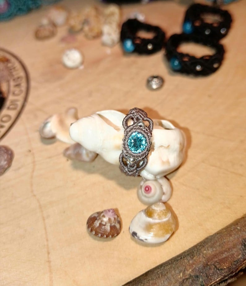 May include: A handmade macrame ring with a light blue gemstone center. The ring is brown and sits on a white seashell, surrounded by other shells and a wooden surface. Other macrame bracelets are in the background.