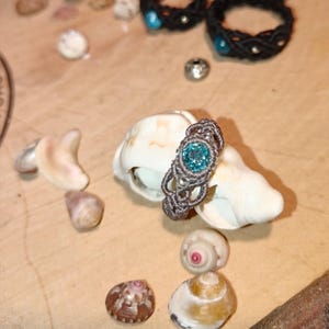 May include: A handmade macrame ring with a teal-colored gemstone set on a seashell, surrounded by other seashells. Several black macrame rings with teal accents are in the background. The ring is a unique piece of jewelry.