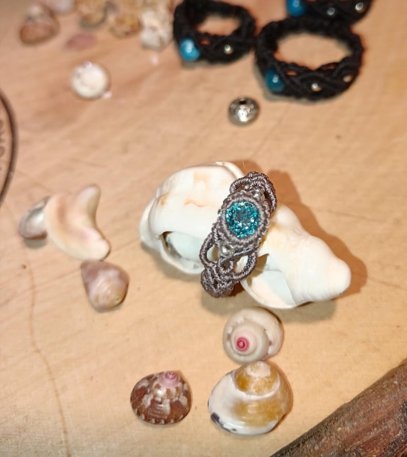May include: A handmade macrame ring with a teal-colored gemstone set on a seashell, surrounded by other seashells. Several black macrame rings with teal accents are in the background. The ring is a unique piece of jewelry.