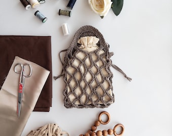 Puede incluir: Una bolsa de red de crochet marrón con un forro de tela blanco. La bolsa está sobre una superficie blanca con varios artículos de costura, incluyendo carretes de hilo, tijeras y una flor blanca.
