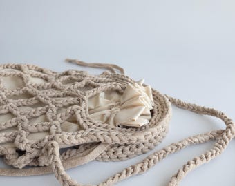 Puede incluir: Una bolsa de crochet beige con cierre de cordón. La bolsa está hecha de hilo grueso tipo cuerda y tiene un tejido suelto y abierto.