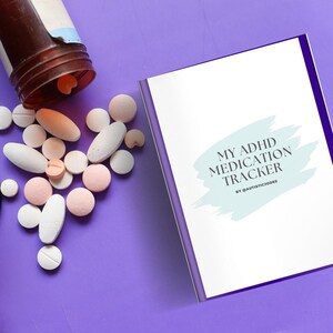 Könnte beinhalten: Eine violette Oberfläche mit einem weißen und blauen Heftchen mit dem Titel "My ADHD Medication Tracker" von @autisticjodes. Um das Heftchen herum liegen weiße und rosa Pillen sowie eine braune Flasche mit weißem Verschluss.