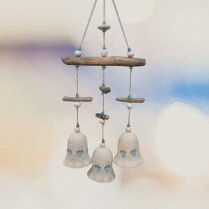 Op de afbeelding: Een hangende mobiel met drie gebroken witte keramische klokjes met hartvormige uitsparingen. De klokjes hangen aan stukjes drijfhout met lichtgekleurd touw, versierd met kleine witte kralen en piepkleine helderblauwe glazen kraaltjes.