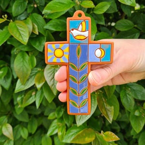 Könnte beinhalten: Ein Keramik-Kreuz mit einem Tauben-, Sonnen- und Mond-Design. Das Kreuz hat einen blauen Hintergrund mit orangefarbenen Rändern und einen zentralen Stiel mit grünen Blättern. Das Kreuz wird vor einem Hintergrund aus grünem Laub gehalten.