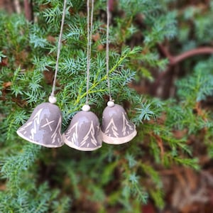 Puede incluir: Tres adornos de campana de cerámica gris con detalles blancos y cuerdas para colgar. Cada campana tiene un diseño único, incluyendo una "X" y patrones de hojas. Las campanas están suspendidas contra un fondo de follaje verde.