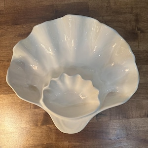 Puede incluir: Plato de servir de cerámica blanca con forma de concha marina con un tazón más pequeño en el centro. El plato tiene un borde rizado y un acabado liso y brillante.