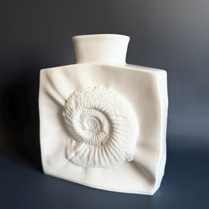 May include: White ceramic vase with a raised, textured ammonite design. The vase has a square shape with rounded edges.