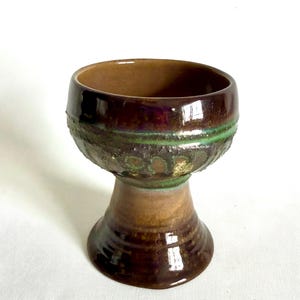 May include: A brown ceramic goblet with a green and gold design. The goblet has a wide bowl and a tall, slender stem.