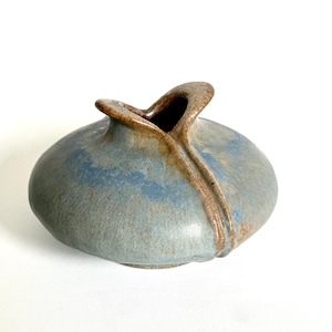 May include: A handmade ceramic vase with a unique shape and a blue-gray glaze. The vase has a rounded body with a flared opening and a brown accent. The vase is approximately 6 inches wide and 4 inches tall.