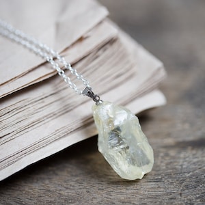 May include: A silver chain necklace with a clear, light yellow crystal pendant. The pendant is a rough, natural shape and is hanging on a stack of old, brown paper.