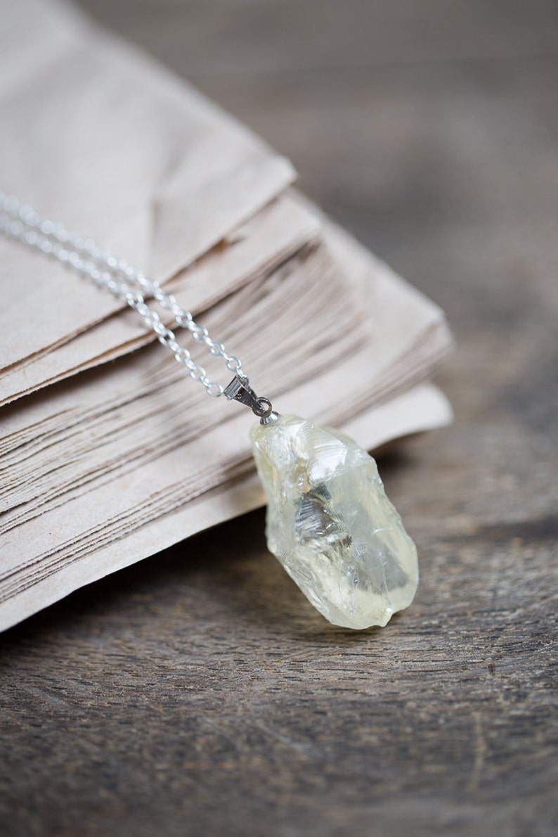 May include: A silver chain necklace with a clear, light yellow crystal pendant. The pendant is a rough, natural shape and is hanging on a stack of old, brown paper.