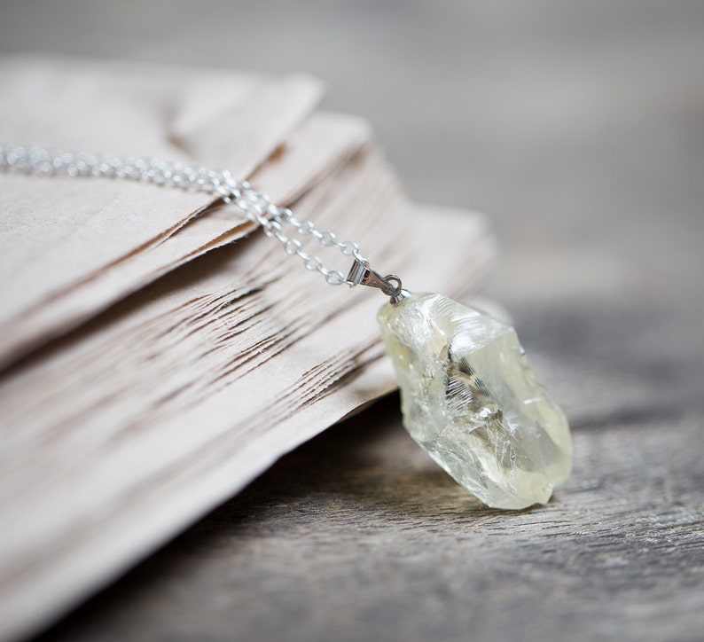 May include: A silver chain necklace with a clear, faceted, yellow crystal pendant. The necklace is lying on a stack of old, cream-coloured paper.