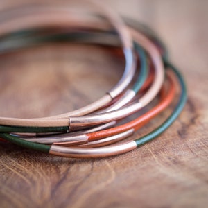 Op de afbeelding: Een stapel leren armbanden met koperen accenten. De armbanden zijn in tinten groen, oranje en beige.
