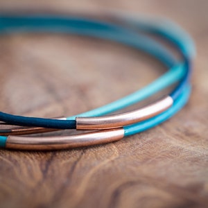 May include: A close-up of a layered leather bracelet with a copper band. The bracelet is made of three different shades of blue leather.