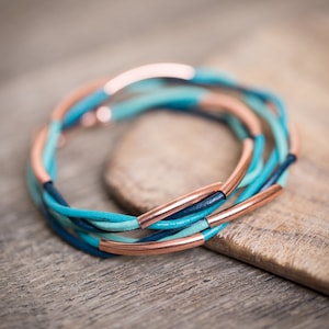 May include: A multi-strand bracelet made of blue and turquoise leather with copper accents. The bracelet is woven in a braided pattern.
