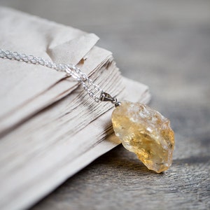 May include: A silver chain necklace with a large, raw, yellow citrine pendant. The necklace is lying on a stack of old, white paper.
