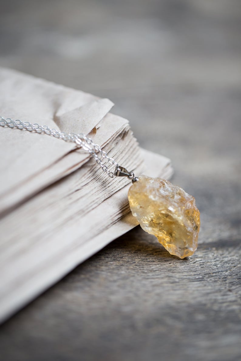 May include: A silver chain necklace with a large, raw, yellow citrine pendant. The necklace is lying on a stack of old, white paper.