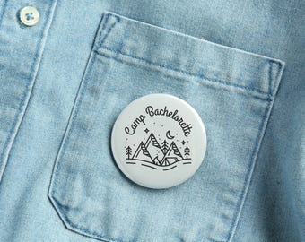 May include: A white button pin with the words "Camp Bachelorette" in a circular design. The pin features a black line drawing of mountains, trees, stars, and a crescent moon. The pin is set against a light blue denim shirt.