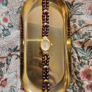 May include: A gold-tone wristwatch featuring an oval face with a white dial and gold hands. The watch band consists of two parallel strands of deep red, round garnet beads connected by small gold-tone metal spacers and a gold-tone clasp.