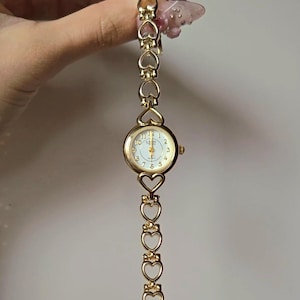 May include: Gold-toned wristwatch with a white face and gold hands. The watch features a round face with numbers and a heart-shaped link band. The band is made of multiple heart-shaped links. The watch is a delicate piece of jewelry.