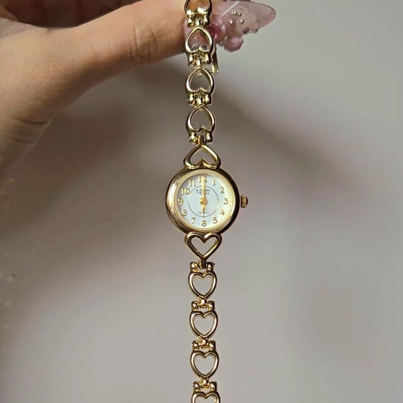 May include: Gold-toned wristwatch with a white face and gold hands. The watch features a round face with numbers and a heart-shaped link band. The band is made of multiple heart-shaped links. The watch is a delicate piece of jewelry.