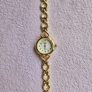 May include: A gold-toned wristwatch with a heart-shaped link band. The watch face is round with a white background, black numerals, and gold hands. The watch is a quartz movement.