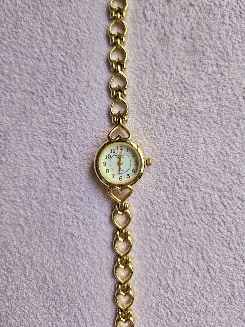May include: A gold-toned wristwatch with a heart-shaped link band. The watch face is round with a white background, black numerals, and gold hands. The watch is a quartz movement.