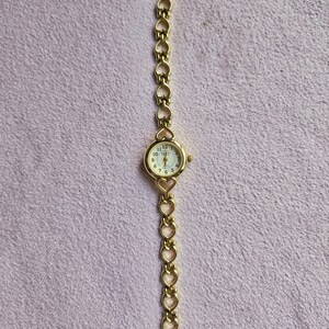 May include: A gold-toned wristwatch with a round face and a heart-shaped link band. The watch face has a white background with black numerals and hands. The band is made of connected heart shapes.