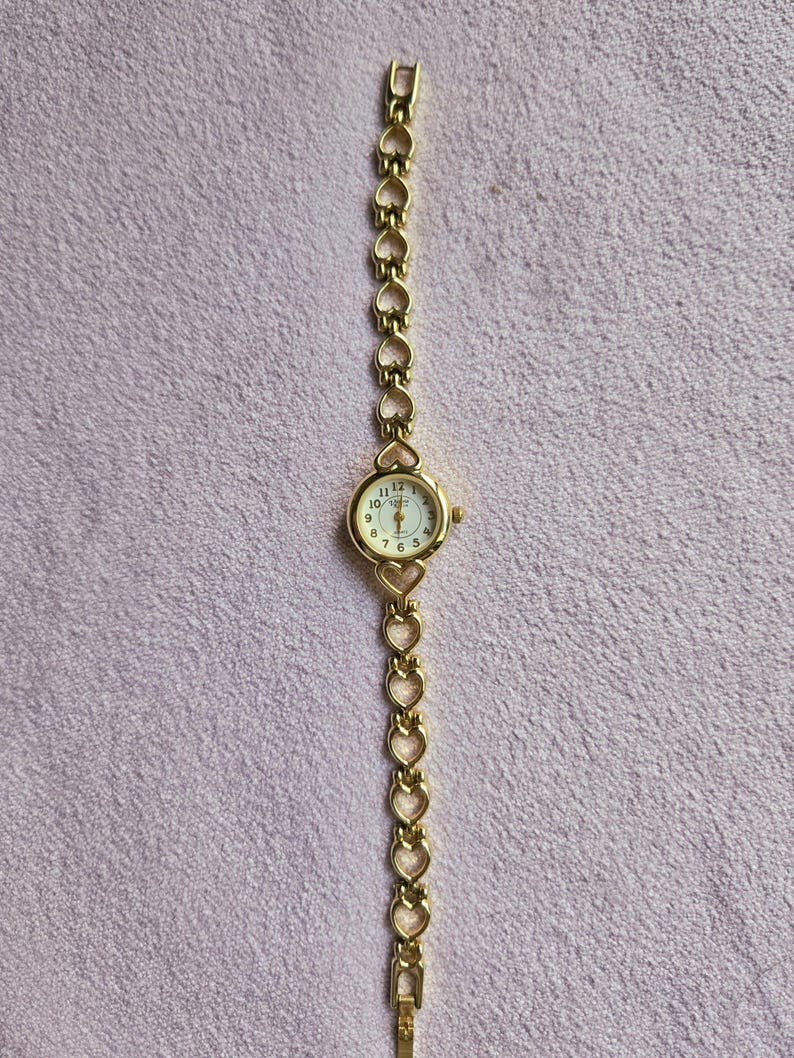 May include: A gold-toned wristwatch with a round face and a heart-shaped link band. The watch face has a white background with black numerals and hands. The band is made of connected heart shapes.