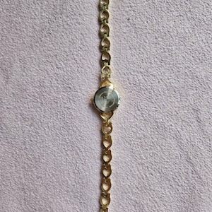 May include: A gold-toned wristwatch with a heart-shaped link band. The watch face is round with a silver-colored dial. The band is connected to the watch face with a small, gold-toned clasp. The watch is lying on a pink surface.