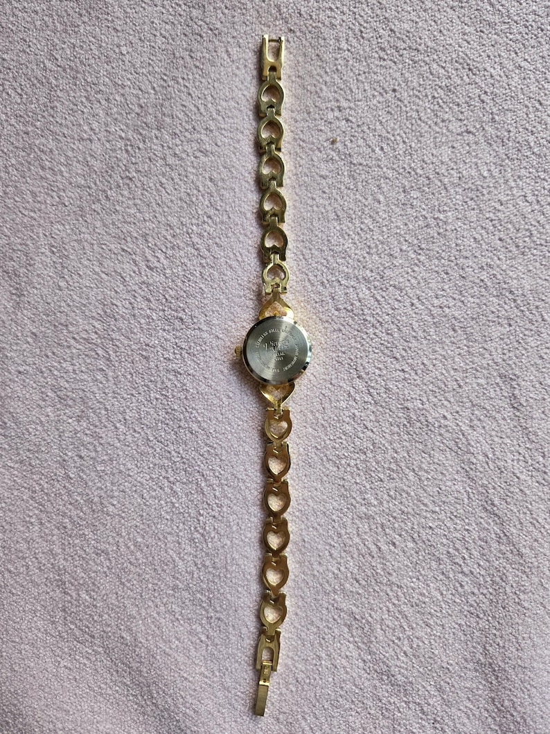May include: A gold-toned wristwatch with a heart-shaped link band. The watch face is round with a silver-colored dial. The band is connected to the watch face with a small, gold-toned clasp. The watch is lying on a pink surface.