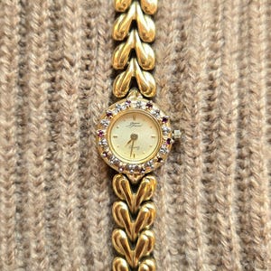 May include: A gold-tone quartz watch featuring a round face with a cream dial, surrounded by a bezel set with alternating small clear stones and red stones. The watch has a distinctive link bracelet composed of repeating heart-shaped gold-tone links.