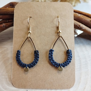 Peut inclure: Une paire de boucles d'oreilles artisanales. Chaque boucle d'oreille présente un cadre en forme de goutte d'eau de couleur or avec un motif tissé bleu marine en bas. Un petit charme rond de couleur or est suspendu au centre de la section tissée.