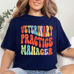 May include: Navy blue t-shirt with the words "VETERINARY PRACTICE MANAGER" in colorful, retro-style lettering. The text is in orange, red, blue, green, and yellow. The shirt is a casual, comfortable style.