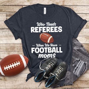 Könnte beinhalten: Ein dunkelblaues T-Shirt mit weißem Text, der "Wer braucht Schiedsrichter - Wenn wir Football-Mütter haben" lautet. Ein brauner und weißer Fußball ist über dem Text abgebildet.