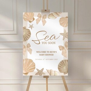 Könnte beinhalten: Willkommensschild für eine Babyparty auf einer Staffelei. Das Schild hat einen beigen Hintergrund mit Illustrationen von Muscheln und Schleifen. Der Text lautet "Sea You Soon" und "Welcome to Beth's Baby Shower". Das Datum ist der 6. September 2025.