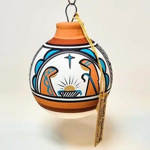 May include: Hand-painted ceramic Christmas ornament with a nativity scene. The ornament features a white base with blue, orange, and black accents. A cross and sunburst design are centered above the nativity figures. A small tag hangs from the ornament.