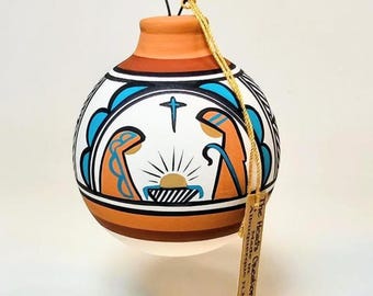 May include: Hand-painted ceramic Christmas ornament with a nativity scene. The ornament features a white base with blue, orange, and black accents. A cross and sunburst design are centered above the nativity figures. A small tag hangs from the ornament.