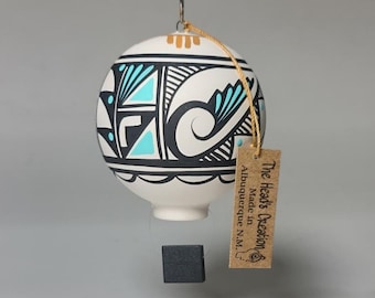 May include: White ceramic ornament with a black and turquoise geometric design. The ornament is hanging from a string and has a small black weight at the bottom. The ornament has a tag that reads "The Head's Creation, Made in Albuquerque NM."