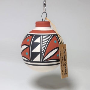 May include: A hand-painted ceramic ornament with a red, white, and black geometric design. The ornament features turquoise accents and a brown tag that reads "The Head's Creation, Made in Albuquerque, N.M."