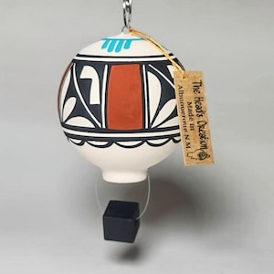 May include: White ceramic hot air balloon ornament with black and red geometric patterns and turquoise accents. The balloon is hanging from a string with a black cube at the bottom. The ornament has a tag that reads "The Headd's Creation, Made in Albuquerque, N.M."