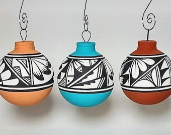 Puede incluir: Tres adornos de cerámica pintados a mano en tonos naranja, turquesa y óxido. Cada adorno presenta un diseño geométrico en blanco y negro y está suspendido por un gancho de alambre. Los adornos son esféricos con un cuello cilíndrico.